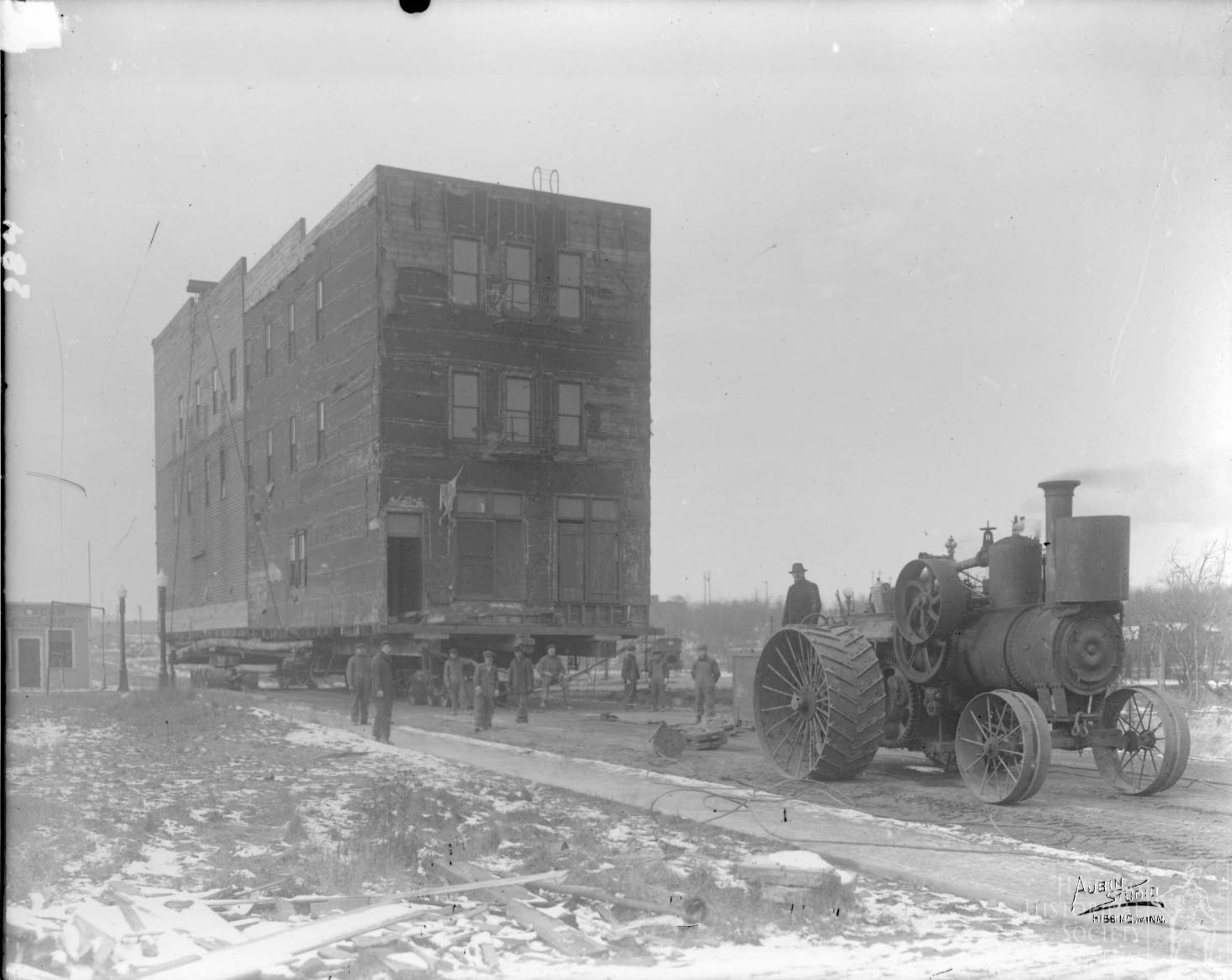 A building moves from north to south Hibbing circa 1918-1922. Starting in 1918, the village of Hibbing was moved two miles south to accommodate the growth of the Hull-Rust-Mahoning mine. Workers jacked up wooden buildings and wheeled them south with steam tractors. Other buildings were scrapped or torn down as the town was rebuilt in its present location.
