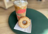 A light, glazed old fashioned doughnut is propped up on a napkin against a tall iced latte with a smiley cloud logo. The whipped cream on top is speckled with black sesam