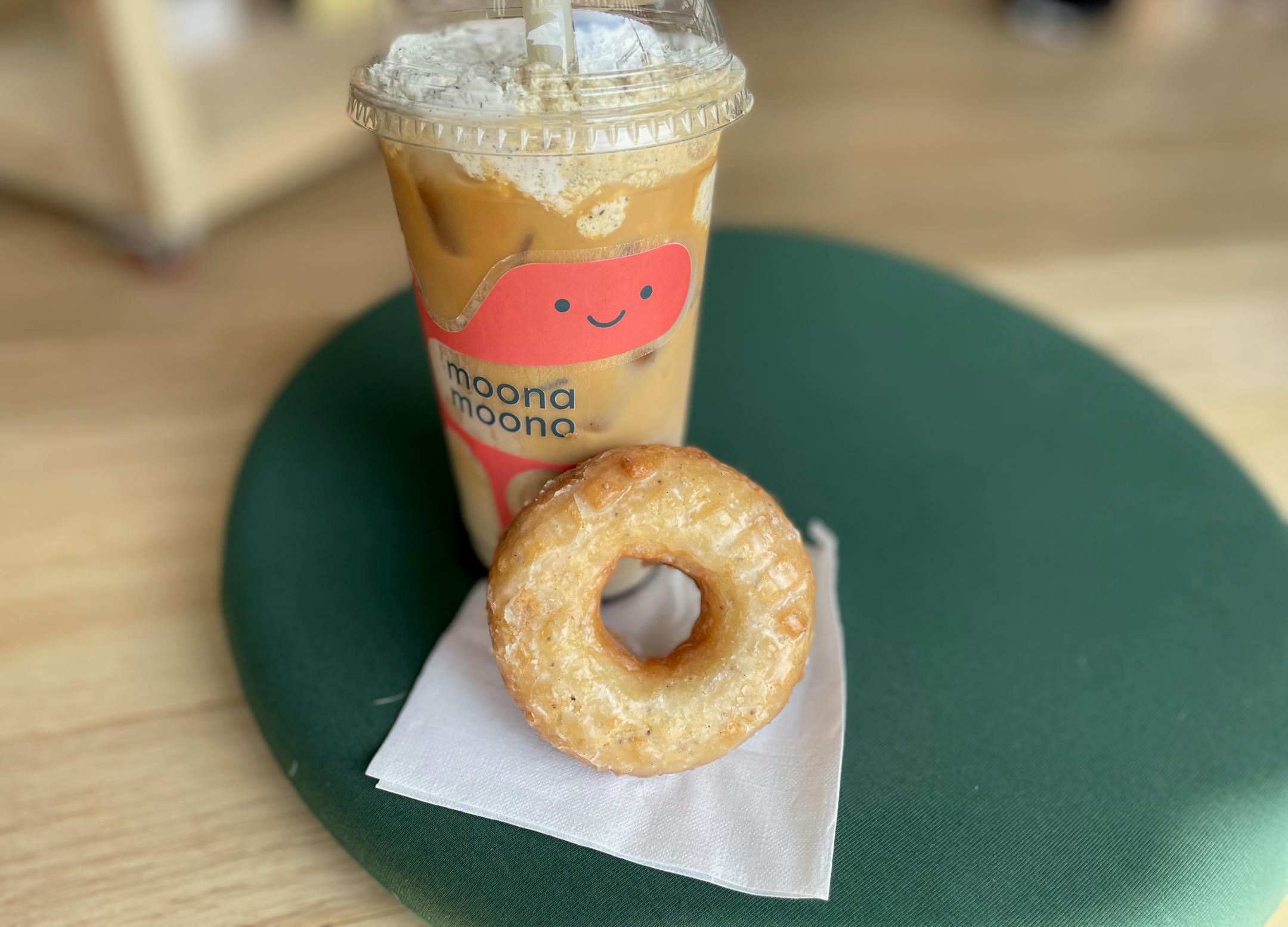 A light, glazed old fashioned doughnut is propped up on a napkin against a tall iced latte with a smiley cloud logo. The whipped cream on top is speckled with black sesam