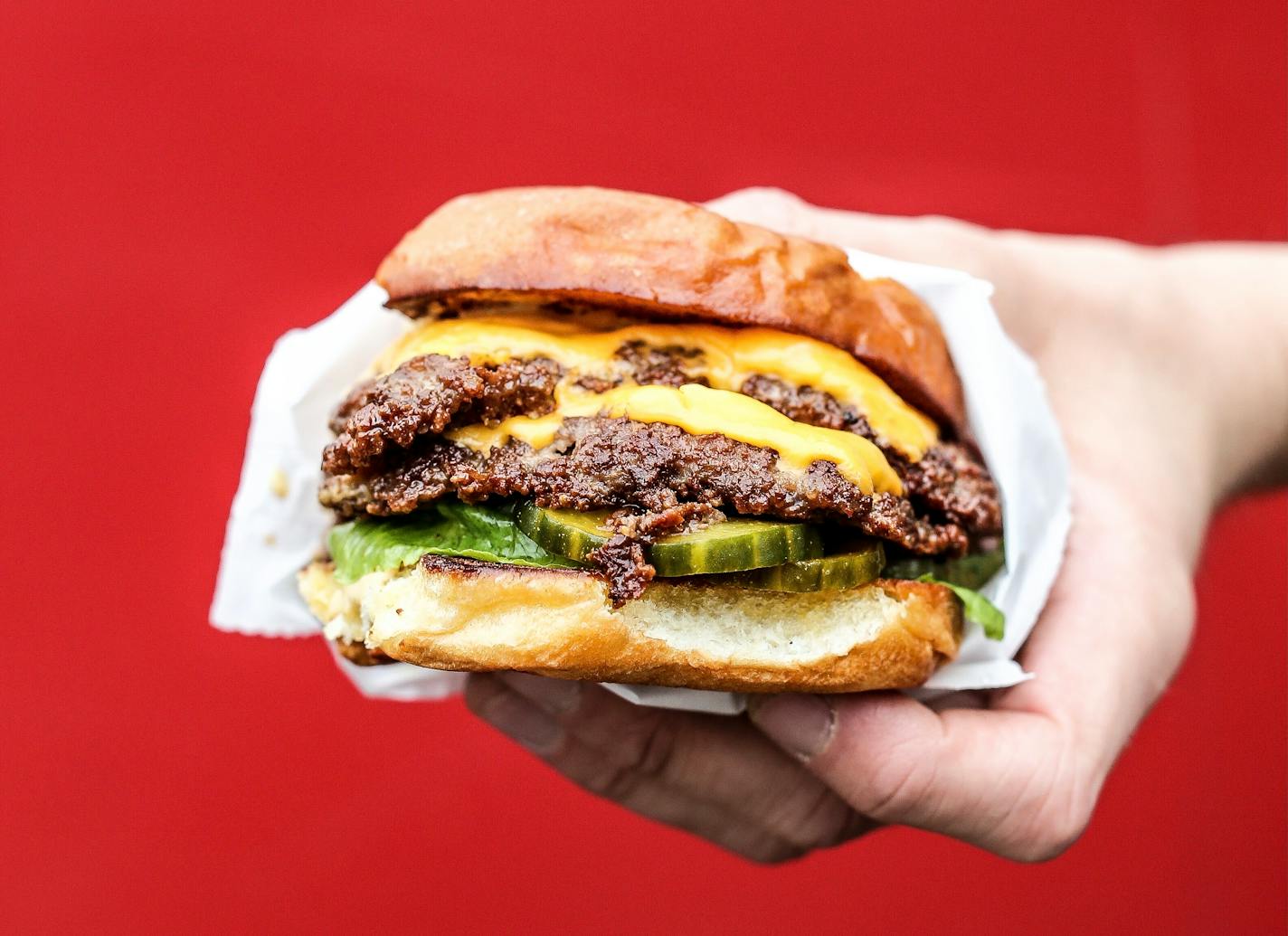 Against a red background, a hand holds a juicy double smashburger with cheese, lettuce ane pickles on a soft bun, wrapped in white wax paper.