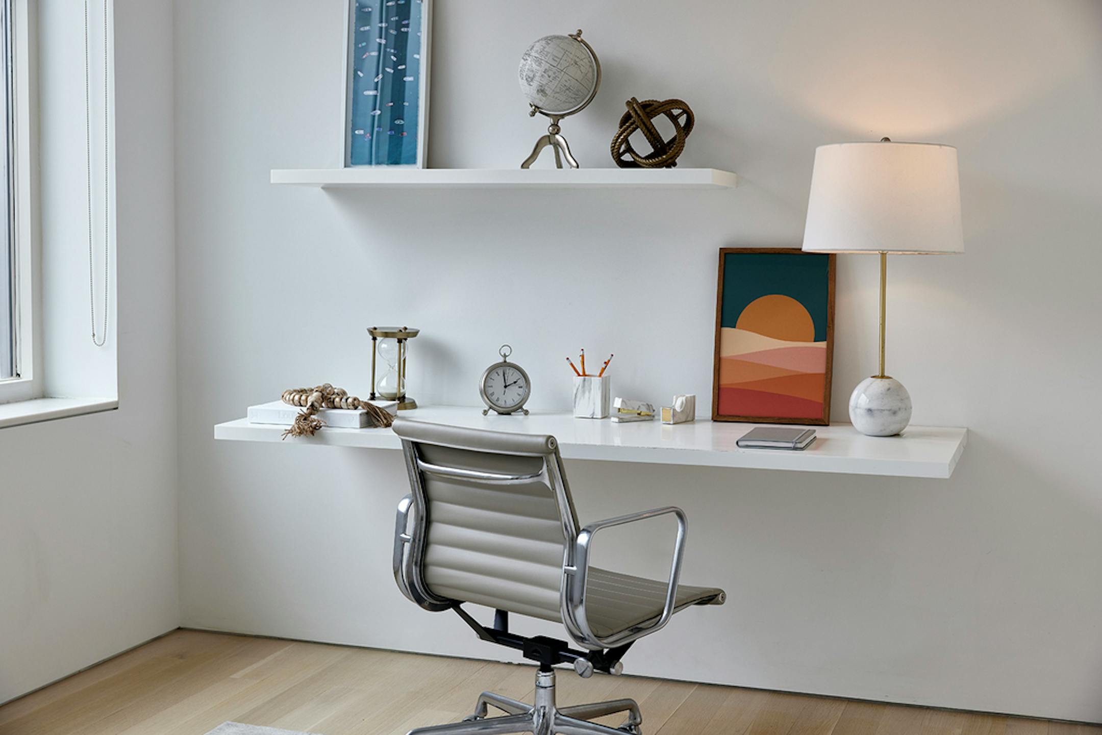 Gray desk chair facing white wall with white shelves, one used as a desk, with framed fabric art.