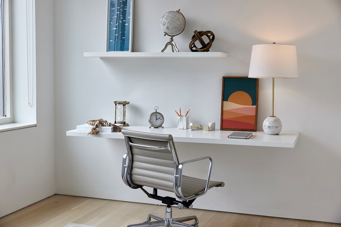 Gray desk chair facing white wall with white shelves, one used as a desk, with framed fabric art.