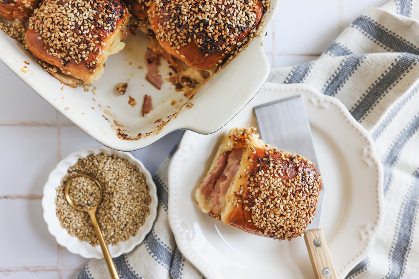 A pan of ham and cheese sliders, topped with sesame seeds and poppy seeds, with a single slider served on a plate and a bowl of sesame seeds on the side.