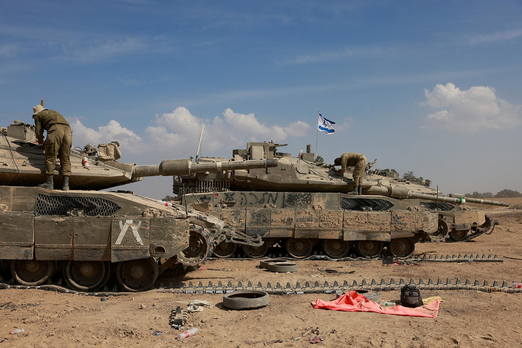 Tanks in a row with solider on top; one has an Israeli flag; on sandy soil with a blue sky with some clouds in the background.
