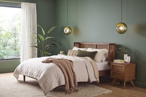 Bedroom with green walls, wood floor and fuzzy pale rug. A window with white curtains is to the left, and there's a green plant, a bed with neutral bedding and a midtone wood end table, with hanging spherical golden lights.