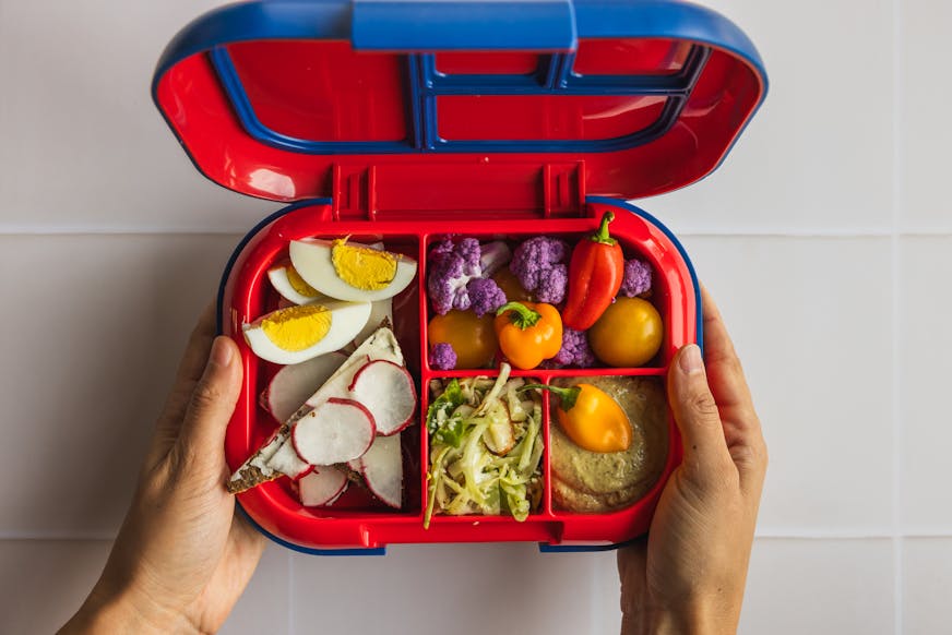 A bright red and blue bento box lunch container with vegetable slaw, hard-cooked eggs and radish points, vegetables and dip.