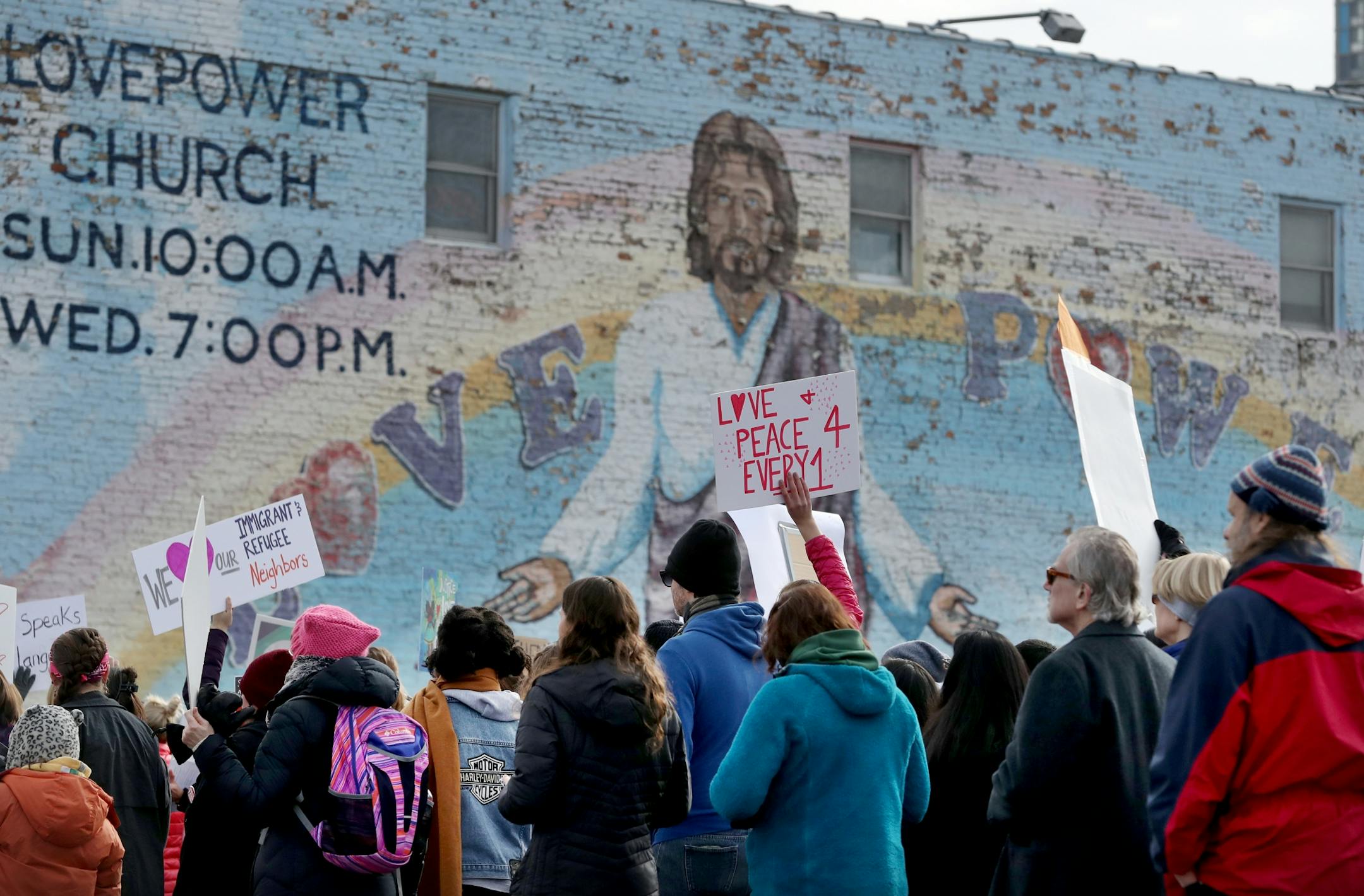 The crowd participating in The Caravan of Love: A Walk of Love for Immigrants and Refugees passes by the famous Love Power Church mural.