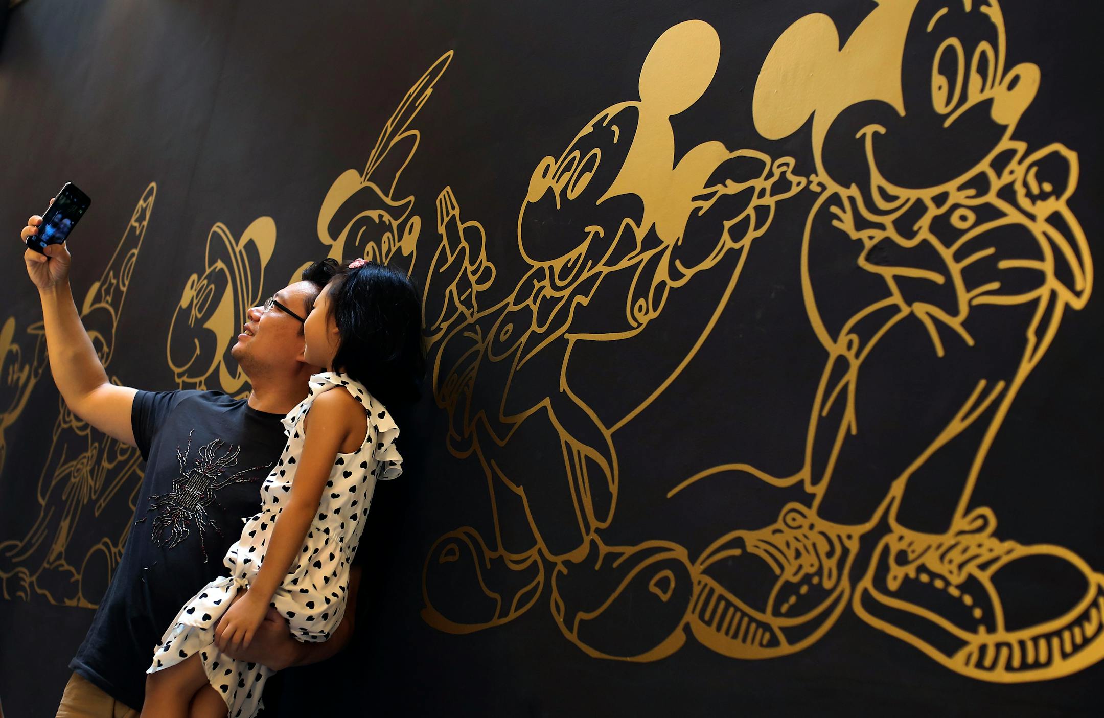 A man holds his daughter as he uses a smartphone to take a selfie in front of a wall with illustrations of Mickey Mouse during the Disney 90th Anniversary Exhibition held in Beijing, China Sunday, Sept. 7, 2014. (AP Photo/Andy Wong)
