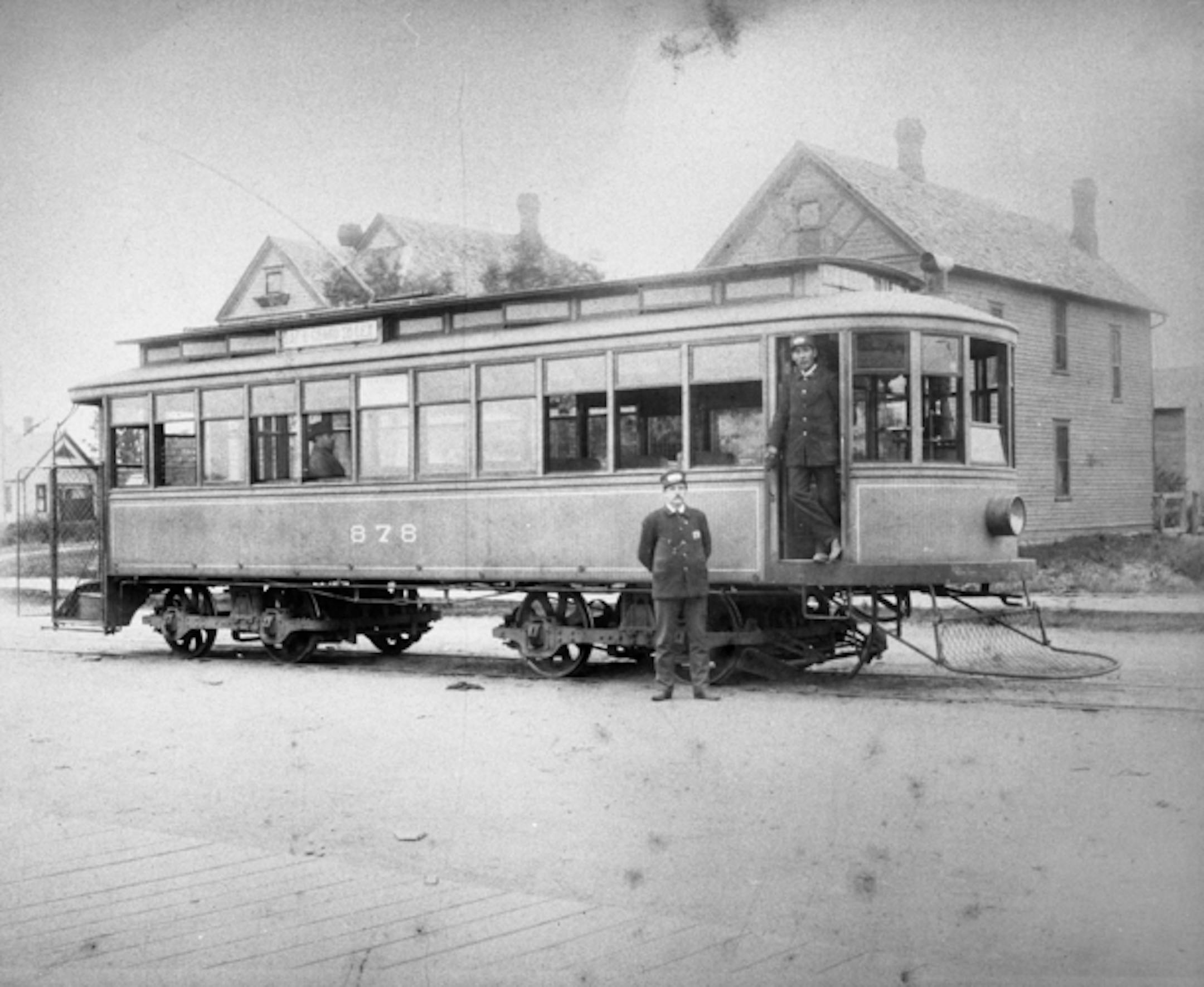 A black-and-white photograph shows a streetcar on Grand Avenue in front of a row of houses. A uniformed man stands in the trolley car's door, and another man in a uniform poses in front of the car.