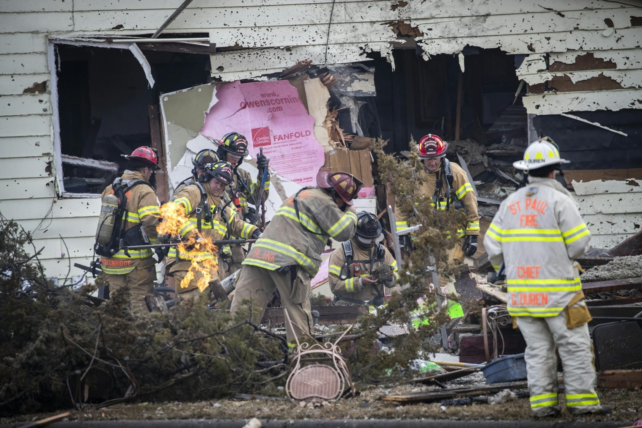 St. Paul firefighters work to clear brush away from a gas line still burning at the scene of a house explosion.
