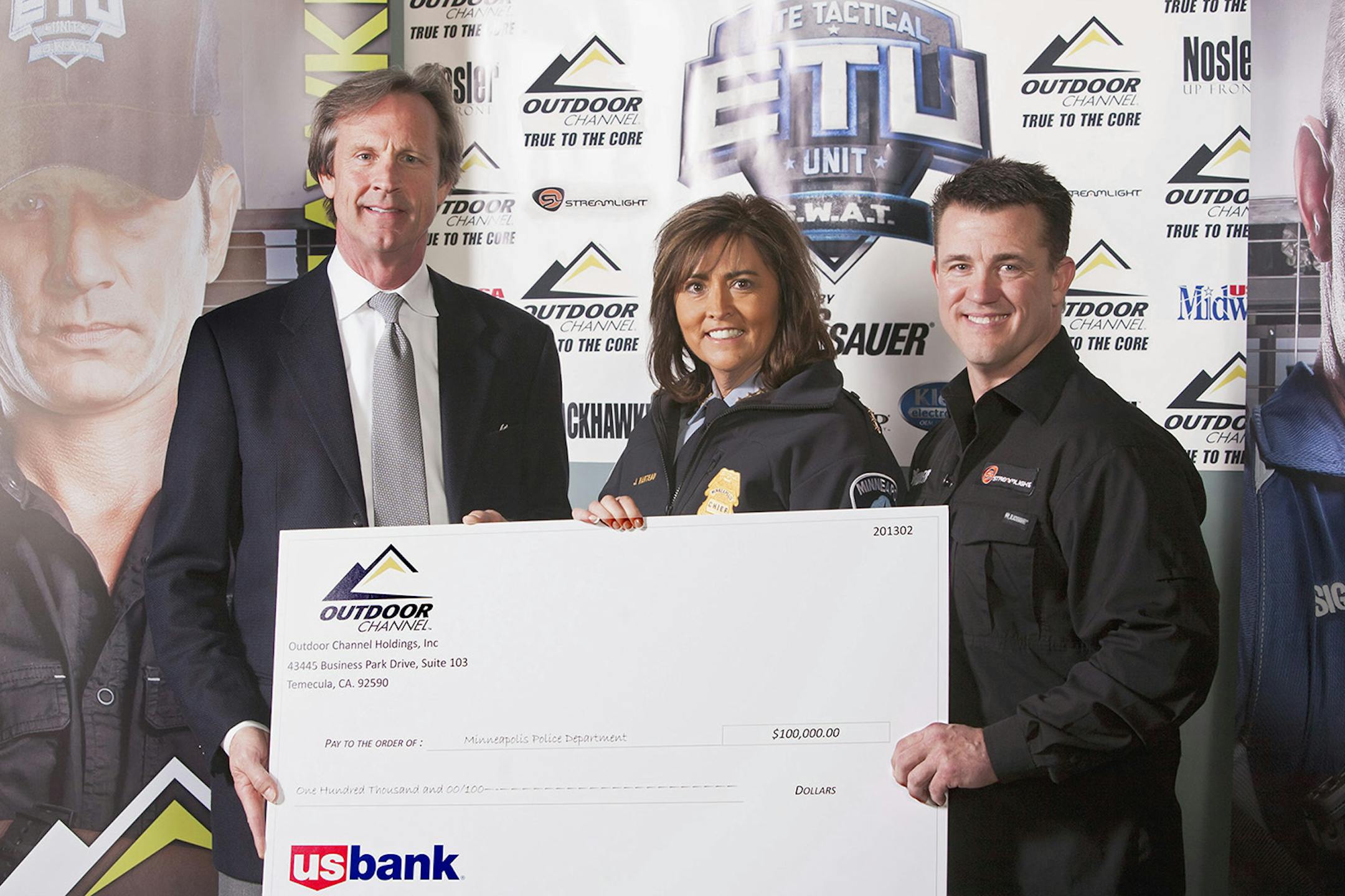 Marc Kidd, Outdoor Channel President of Media Sales, on the left, presenting a mock check of $100,000, to Minneapolis Police Chief Janee Harteau, who is in the center, and Officer Tony Caspers, on the right. There is an accent mark over the first 'e' in Janee