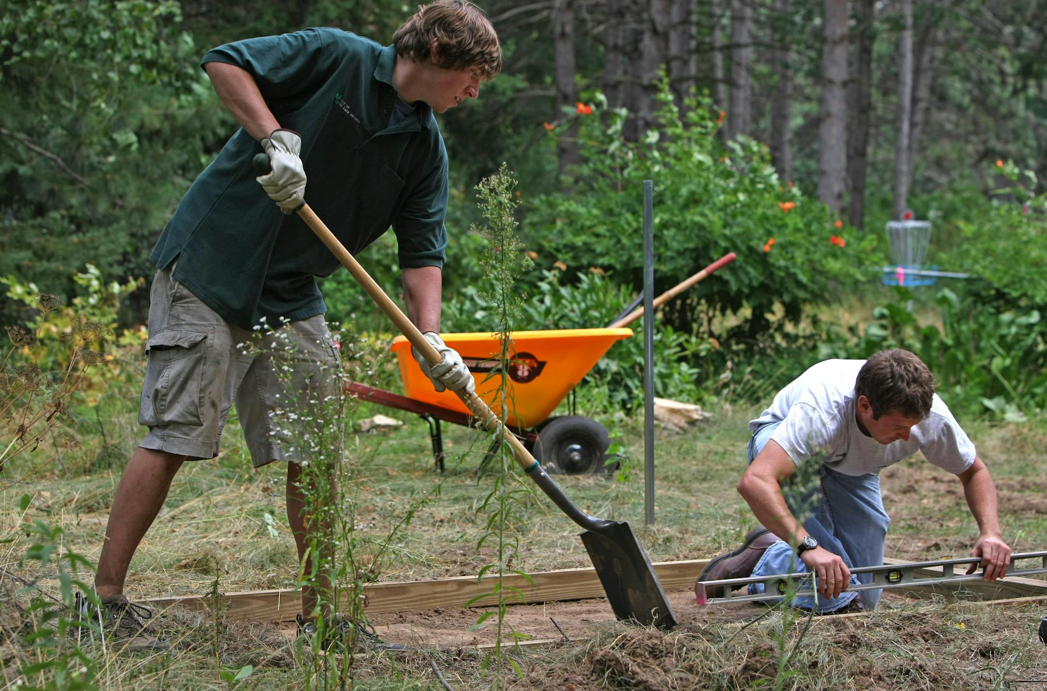 (left to right) Oak Park Heights Intern Eric Eaton and Andy Kegley of the public works department, leveled and graded one of the tee pads on the new disc golf course at Brekke Park in Oak Park Heights. Behind them was one of the course baskets to catch the flying discs.