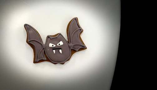 A bat cookie on a white table. Its face is a little frown and it has white frosting fangs