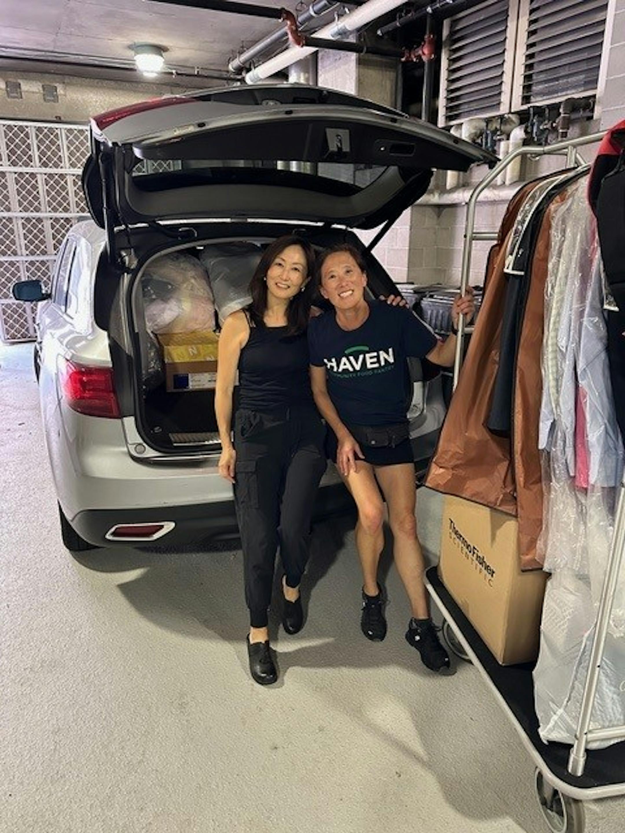 Two Asian women smile in front of a van loaded with bags and boxes. A rolling garment rack is filled with clothing.