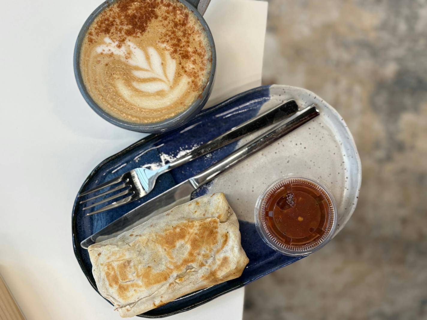 A latte with a sprinkle of cinnamon and floral latte art is in a large, stoneware cup next to a matching plate holding a griddled breakfast burrito and a take out container of salsa.