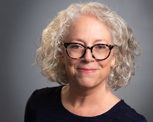 Head shot against a gray background of a woman with gray wavy hear and glasses.