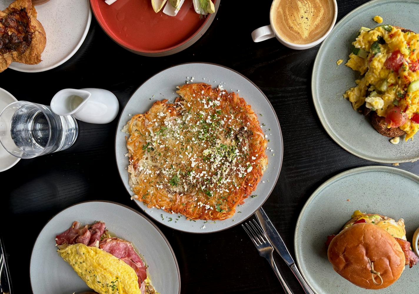 A selection of Altera's brunch items, shot overhead with an egg sandwich, giant hash brown, omelette, scrambled eggs on toast, a latte colored golden with tumeric and a salad.