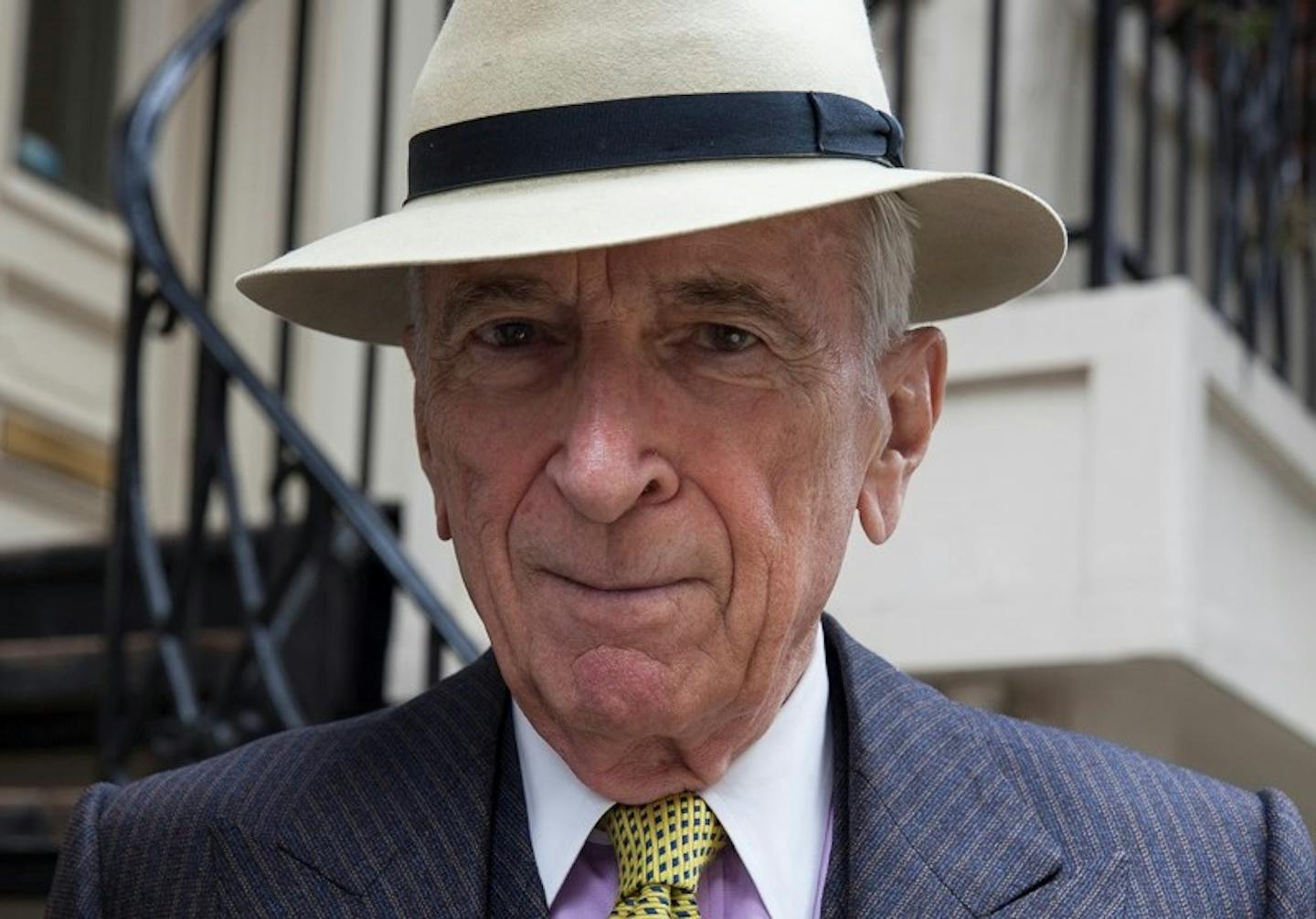 Portrait of Gay Talese wearing white fedora with black ribbon and charcoal pinstripe suit with lavender shirt and yellow and blue tie.