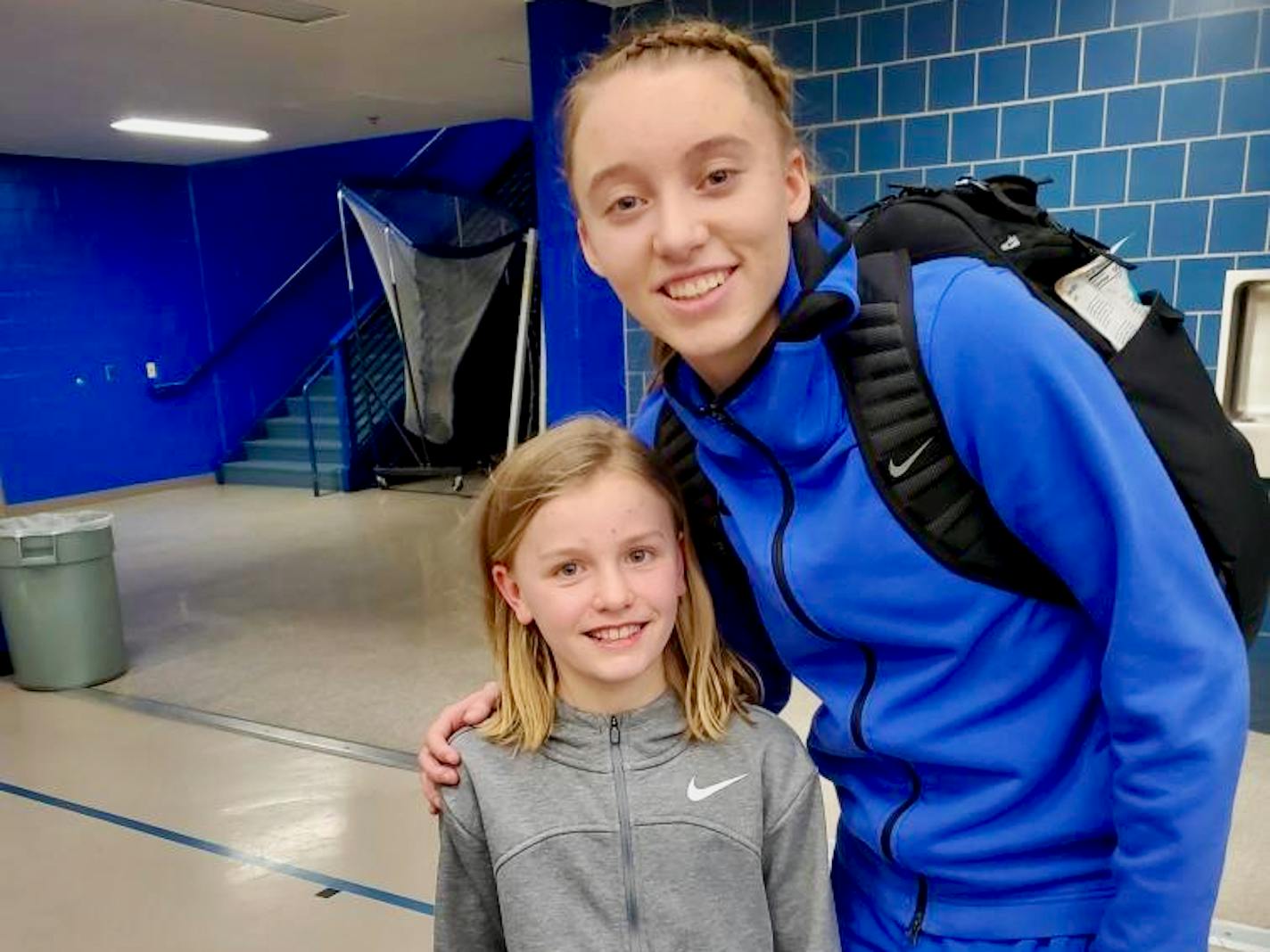 A blond girl with shoulder-length hair wearing a gray Nike track jacket poses next to a blond college athlete wearing a blue track suit