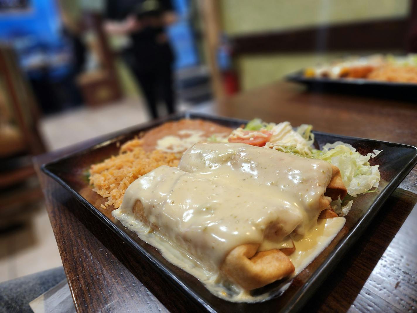 A rectangular plate of cheese-covered chimichangas with rice and beans on a table