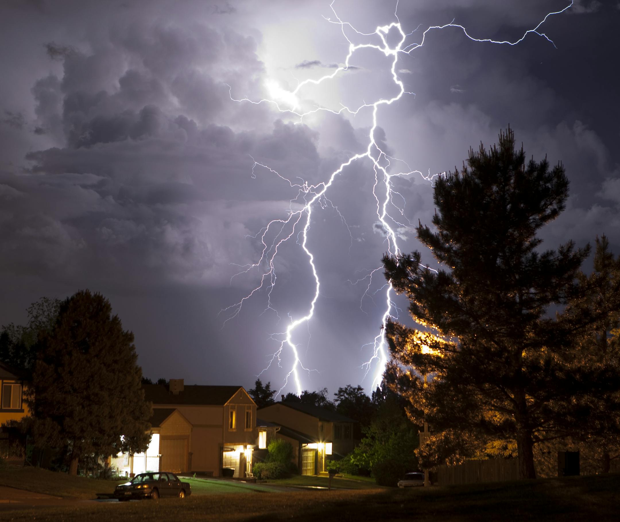 A massive thunderhead pours rain and lightning over suburban Denver homes, Colorado. Trees are silhouetted and homes are lit by incandescent light. The thunderhead is lit by huge lightning bolts reaching from the top of the clouds to the ground.
