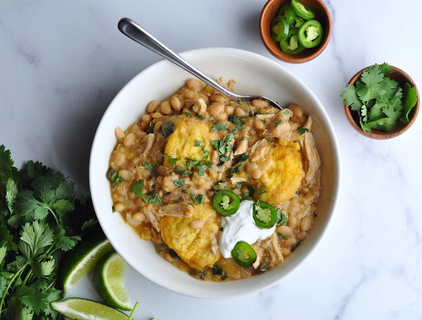 A bowl of white chicken chili and cornbread dumplings garnished with sour cream, jalapeños, cilantro and lime wedges.