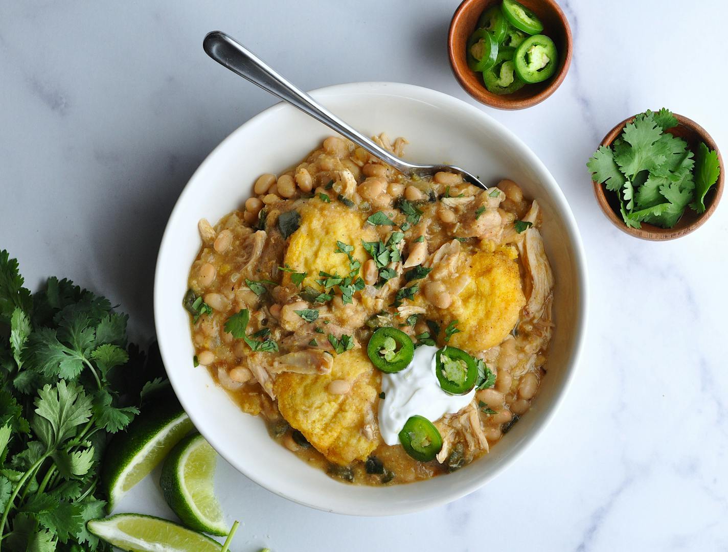 A bowl of white chicken chili and cornbread dumplings garnished with sour cream, jalapeños, cilantro and lime wedges.