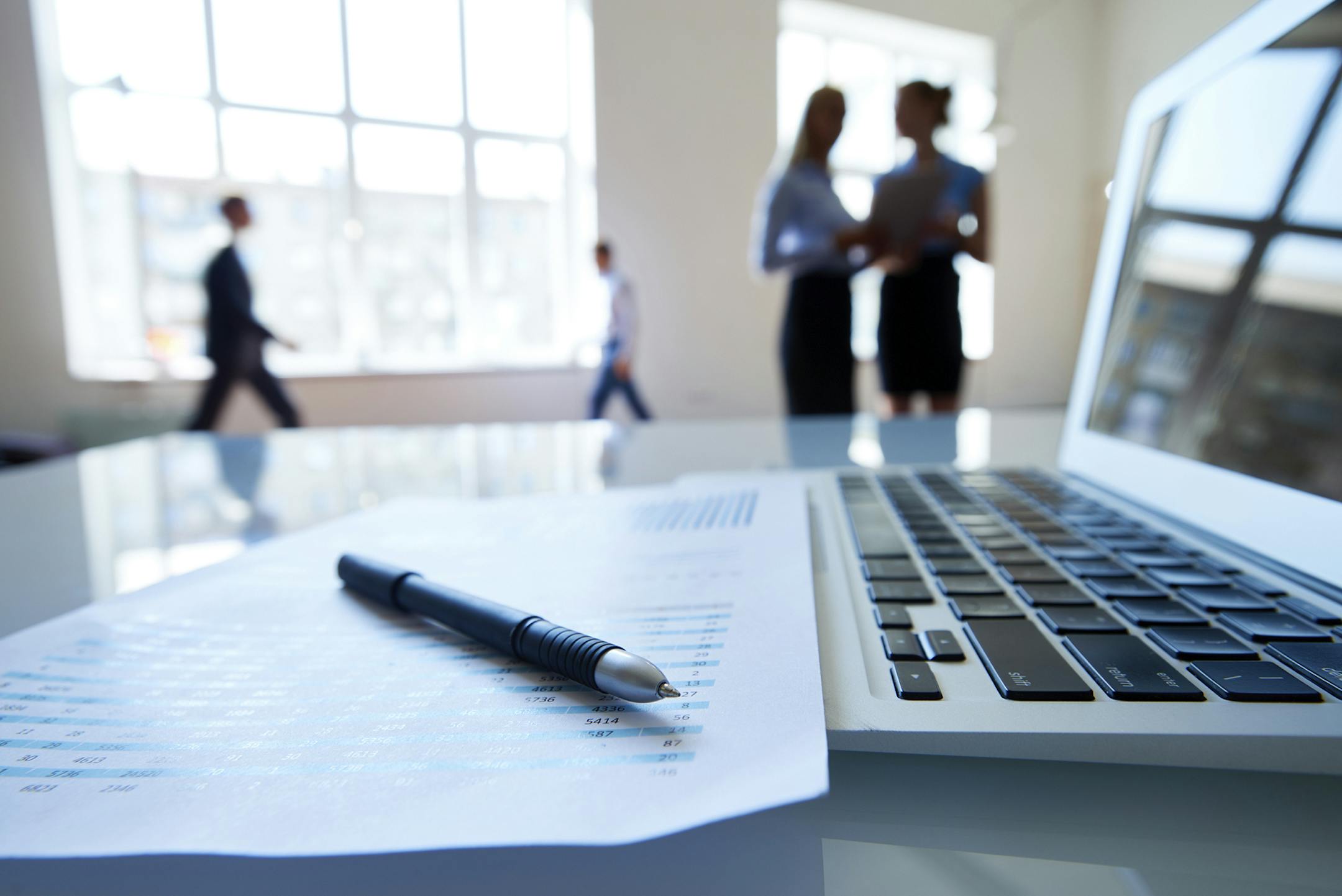 Document with statistical data, laptop and pen on desk, business people in the background