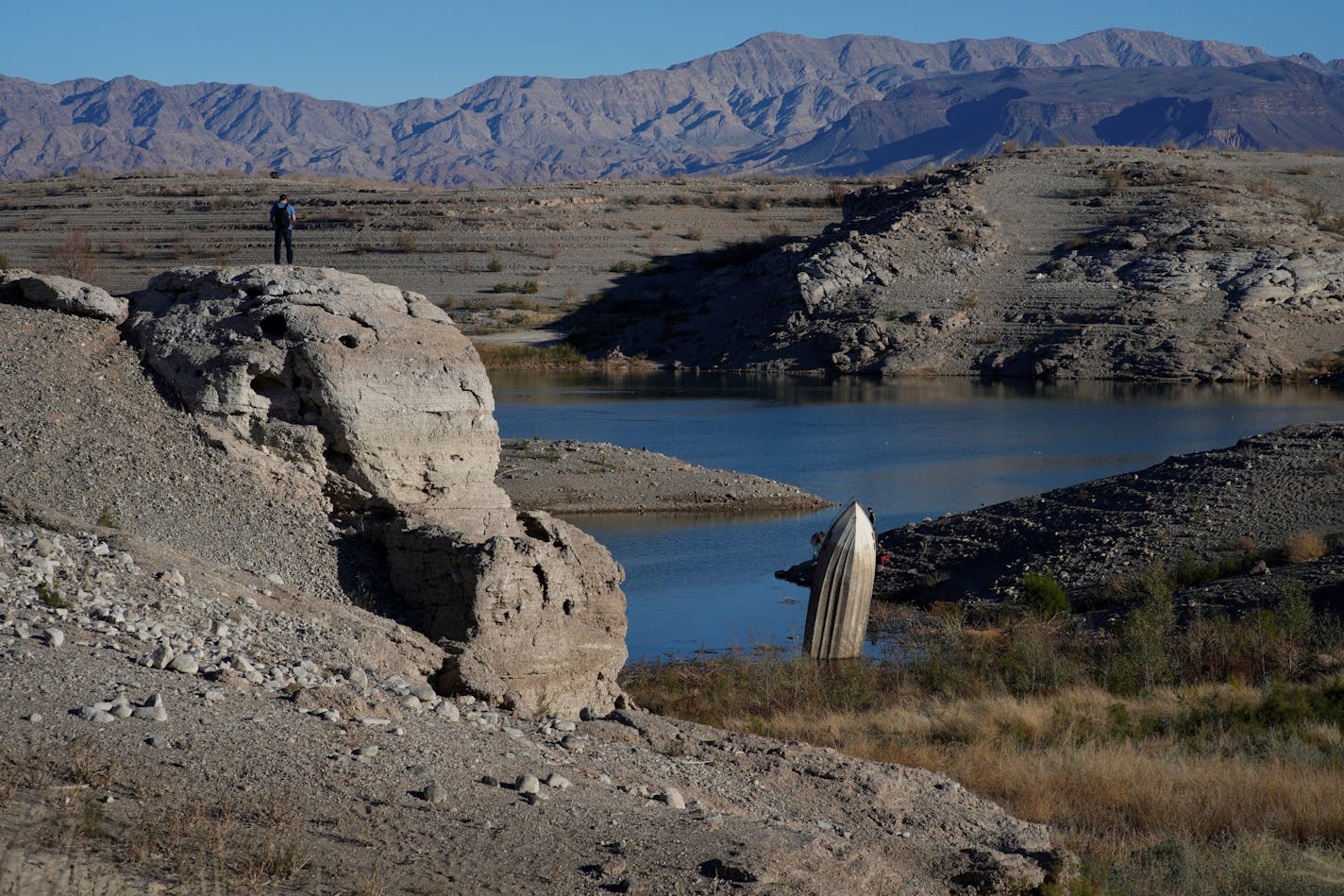 A man stands on a hill overlooking a formerly sunken boat standing upright into the air with its stern buried in the mud along the shoreline of Lake Mead at the Lake Mead National Recreation Area, Friday, Jan. 27, 2023, near Boulder City, Nev. Credit: AP Photo/John Locher