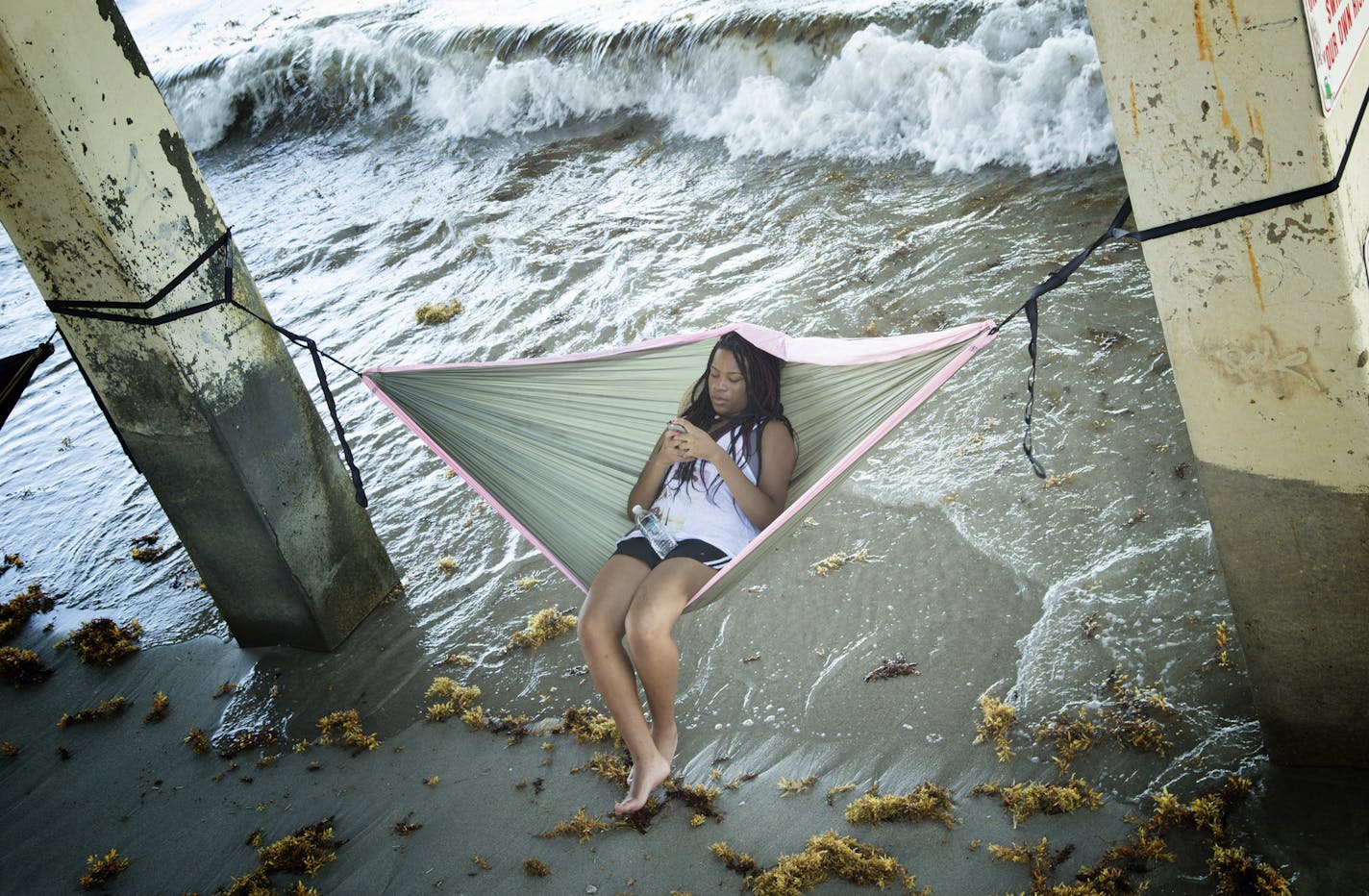 A young girl sits on a hammock under a pier as she plays with her phone at Dania Beach, Fla.