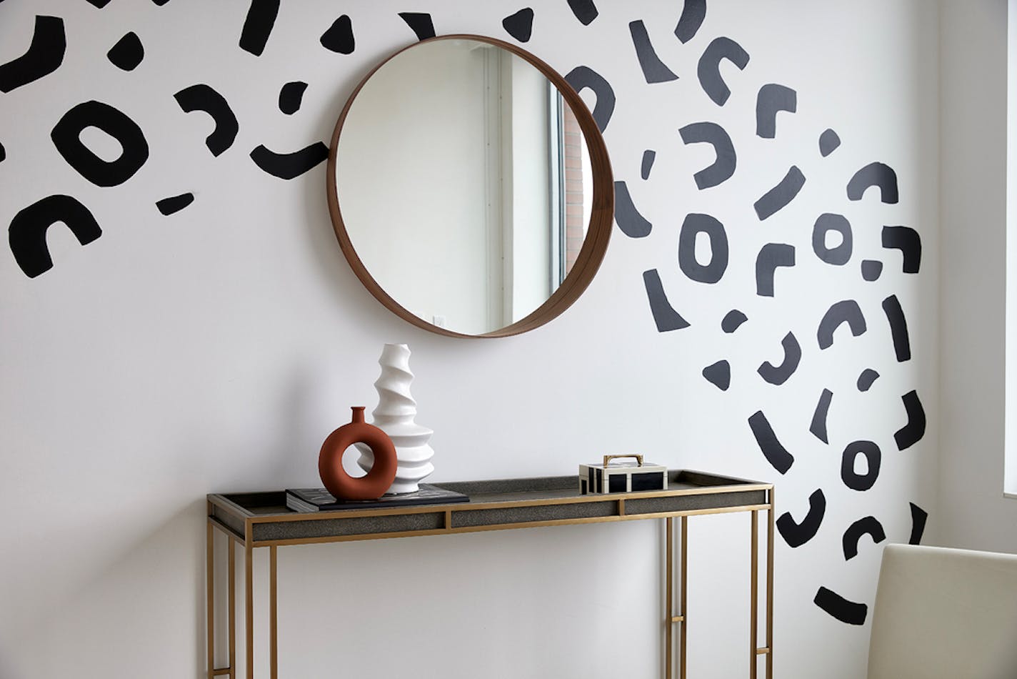 Black cutouts in arc shape on white wall with brass-framed mirror over brass-accented table.