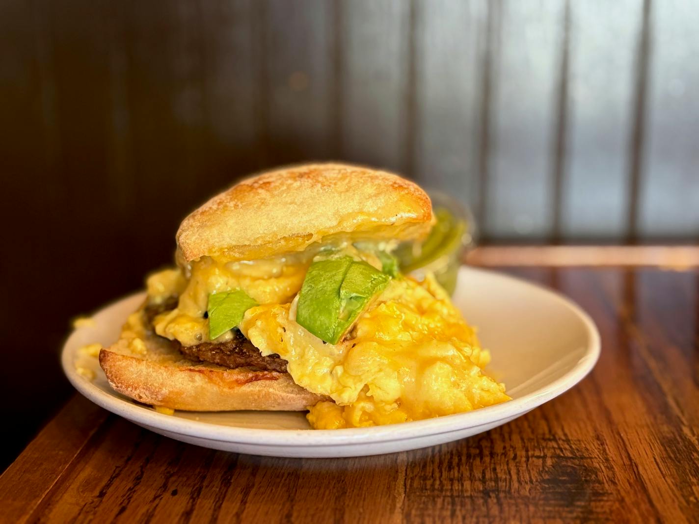 An egg sandwich with soft scrambled eggs, cheese, avocado on ciabatta. Photographed in the coffee shop on a small table with a wood panel background.