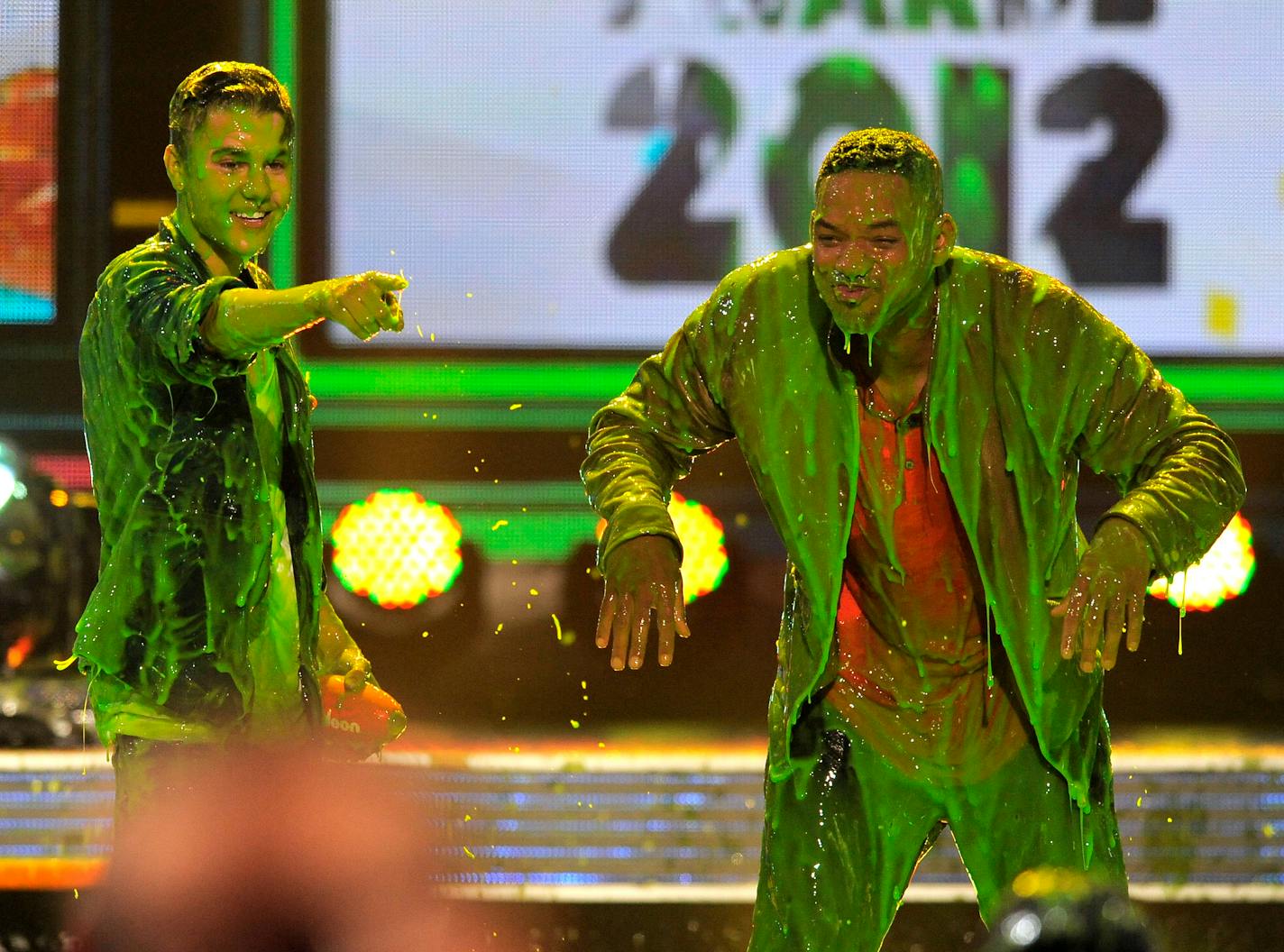 Justin Bieber stands to the left of Will Smith and points off-camera. The two men are drenched in a green slimy liquid.