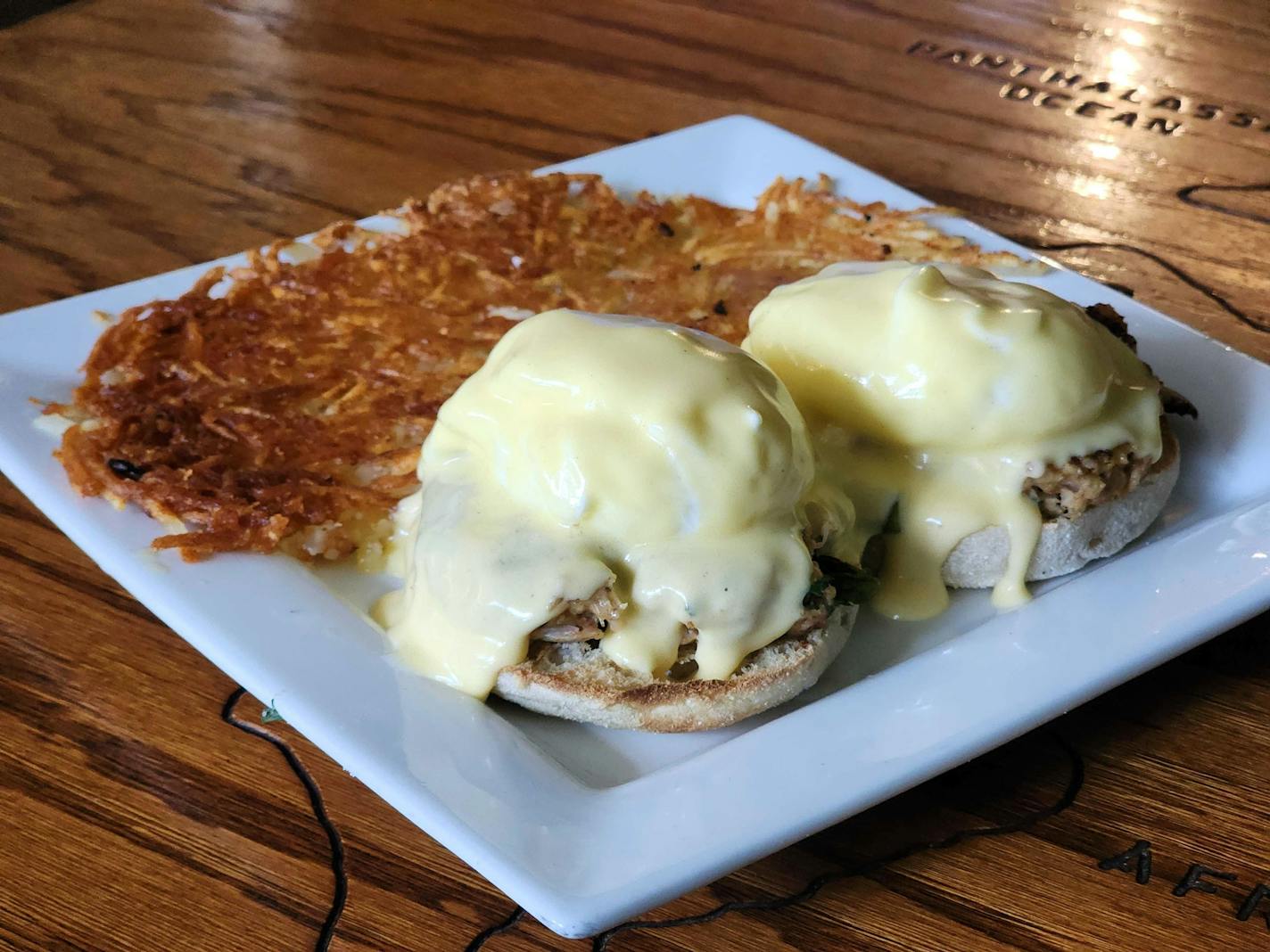 A heavily grained wooden table with map carvings in it, on top of which lays a square white plate with an open-faced English muffin topped with crab cakes, poached eggs, and yellow hollandaise sauce dripping down