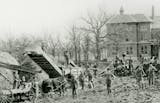 Black and white photographic print of a threshing scene at the Goodhue County Poor Farm. The image depicts a groupd of people standing with horses, wagons, and farm equipment with hay on the left.