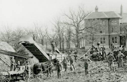 Black and white photographic print of a threshing scene at the Goodhue County Poor Farm. The image depicts a groupd of people standing with horses, wagons, and farm equipment with hay on the left.