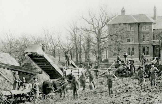 Black and white photographic print of a threshing scene at the Goodhue County Poor Farm. The image depicts a groupd of people standing with horses, wagons, and farm equipment with hay on the left.