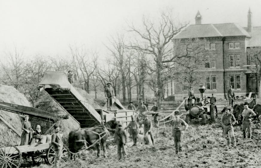 Black and white photographic print of a threshing scene at the Goodhue County Poor Farm. The image depicts a groupd of people standing with horses, wagons, and farm equipment with hay on the left.