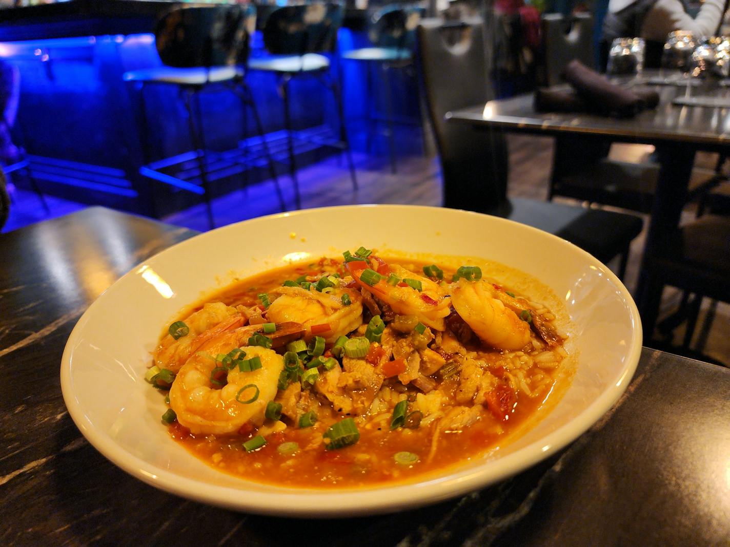 A white bowl containing red stew with pieces of shrimp and chicken, set on a black table in front of a blue glowing light from the bar