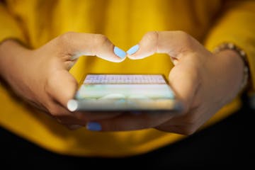 Young woman using cell phone to send text message on social network at night. Closeup of hands with computer laptop in background