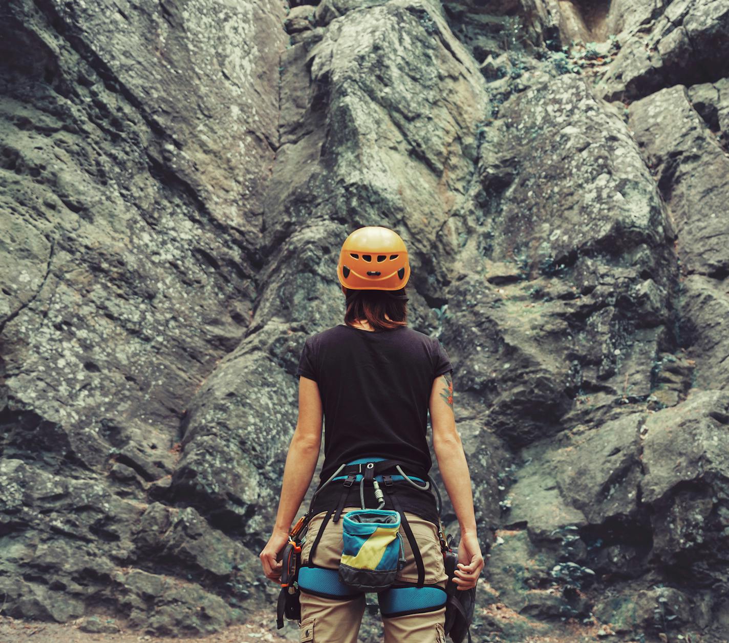 Young woman wearing in climbing equipment standing in front of a stone rock outdoor and preparing to climb, rear view