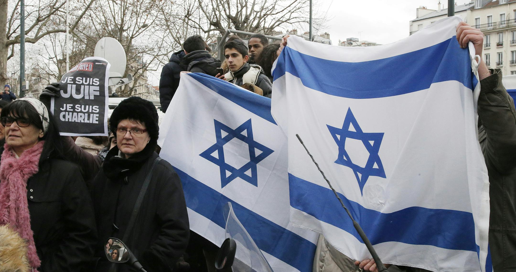People wave Israeli flags after Israeli Prime Minister Benjamin Netanyahu's visit to the kosher market where four hostages were killed, in Paris, France, Monday, Jan. 12, 2015. France's defense minister says the country is mobilizing 10,000 security forces to protect the country after three days of terror attacks. French Interior Minister Bernard Cazeneuve said 4,700 security forces would be assigned to protect France's 717 Jewish schools. Poster at left reads: "I am Jewish, I am Charlie". (AP P