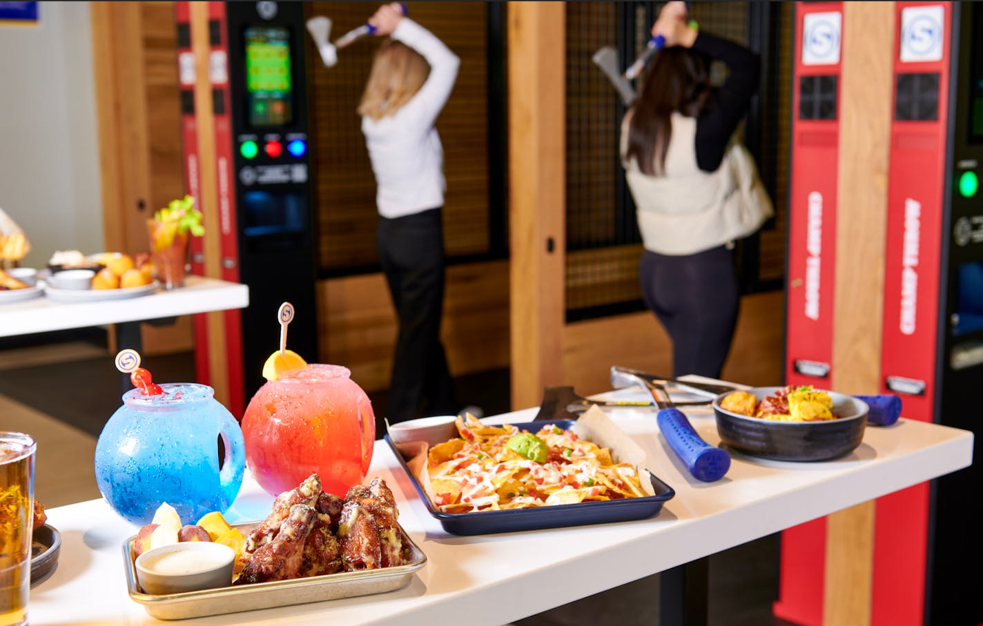 Two women can be seen getting ready to throw their axes. In the foreground, appetizers  including chicken wings and nachos are displayed on a table beside colorful cocktails in fishbowl-like mugs.