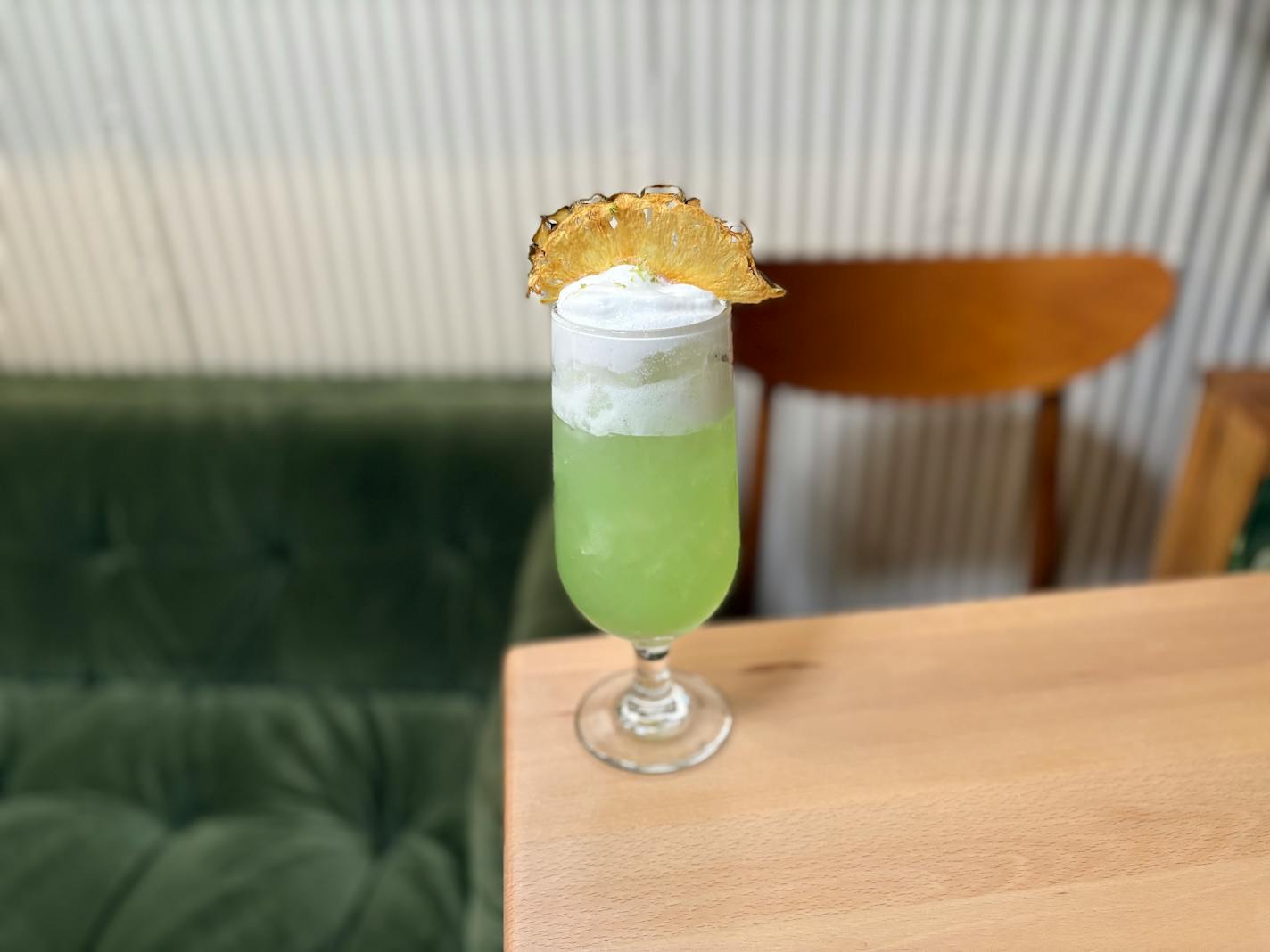 A vibrant, light green colored cocktail with a thick, white foam topping and dehydrated pineapple garnish.