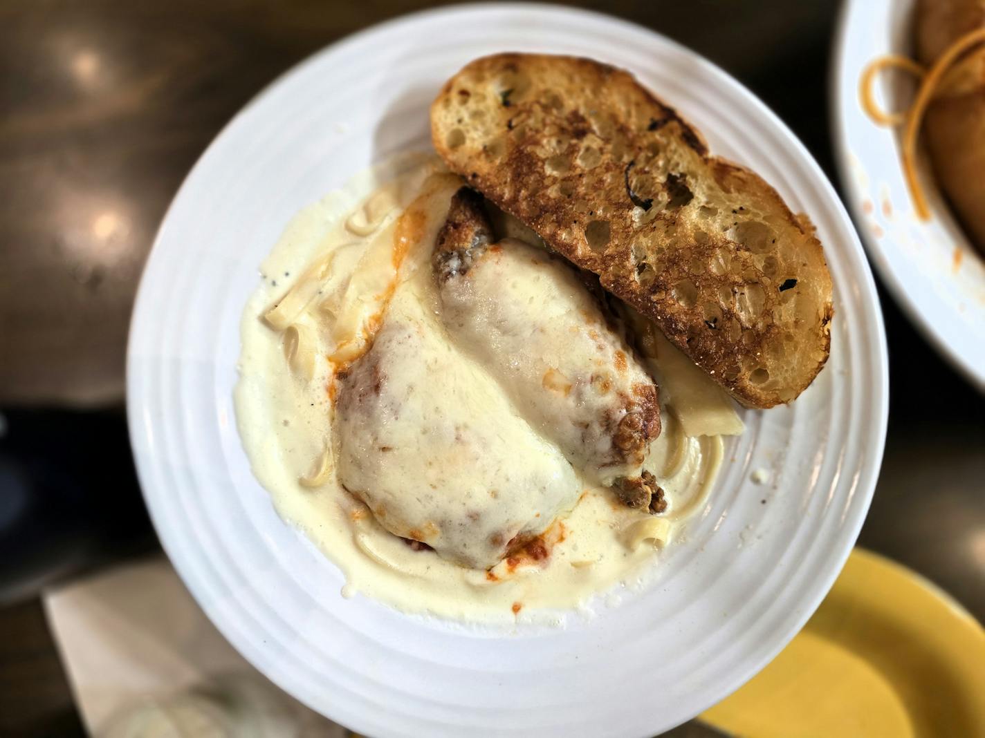 A white bowl with a pool of white alfredo sauce and two cheese-covered fried chicken breast pieces, and a slab of garlic toast.