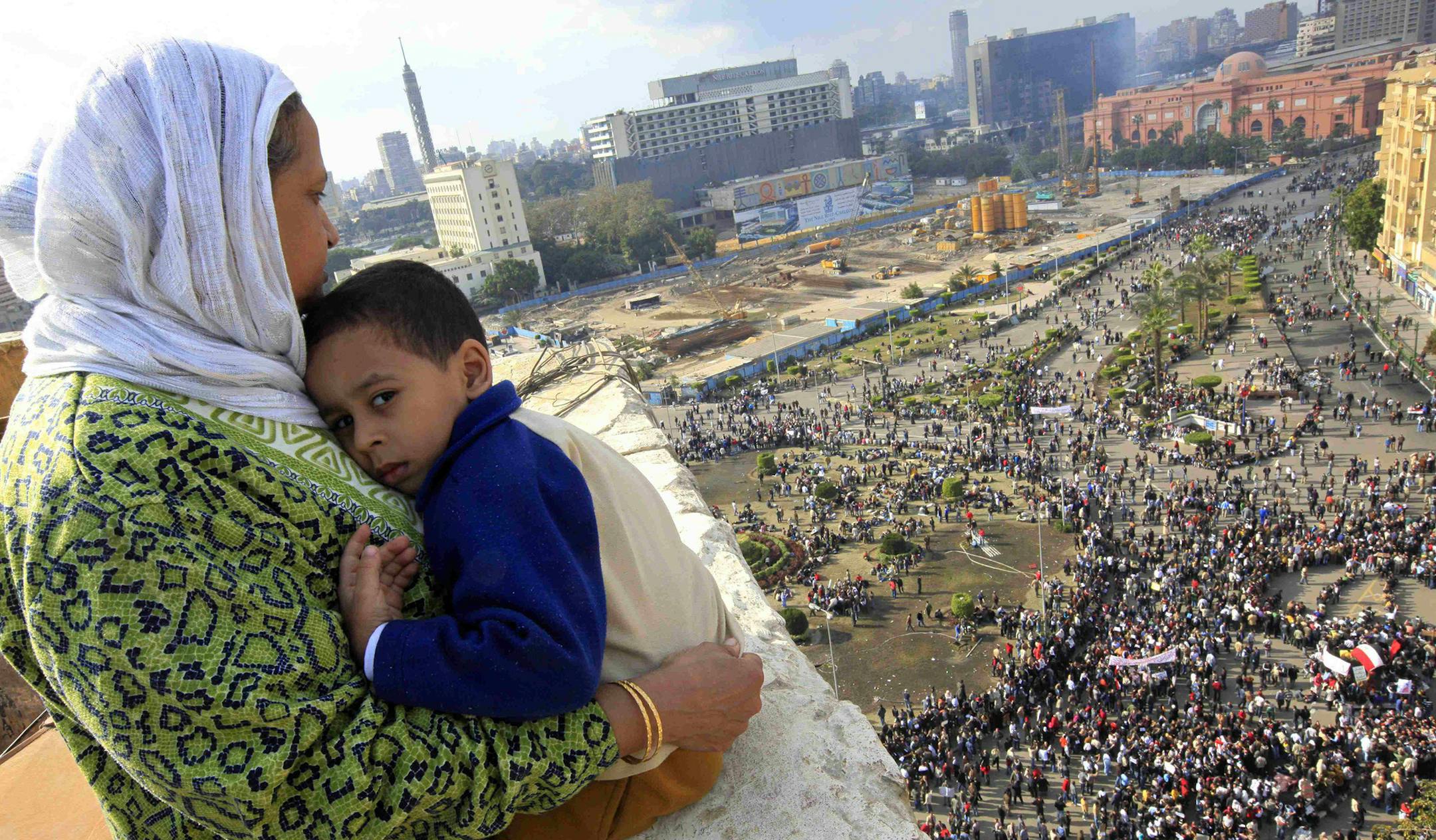 An Egyptian mother hugs her child as she watches some thousands of Egyptian protesters gather at Tahrir square in Cairo, Egypt, Sunday, Jan. 30, 2011, with the ruling National Democratic party building burned at top right behind the red coloured Egypt museum. The army sent hundreds more troops and armored vehicles onto the streets of Cairo and other cities but appeared to be taking little action against mass protests on Sunday. (AP Photo/Amr Nabil) ORG XMIT: MIN2016012210435988