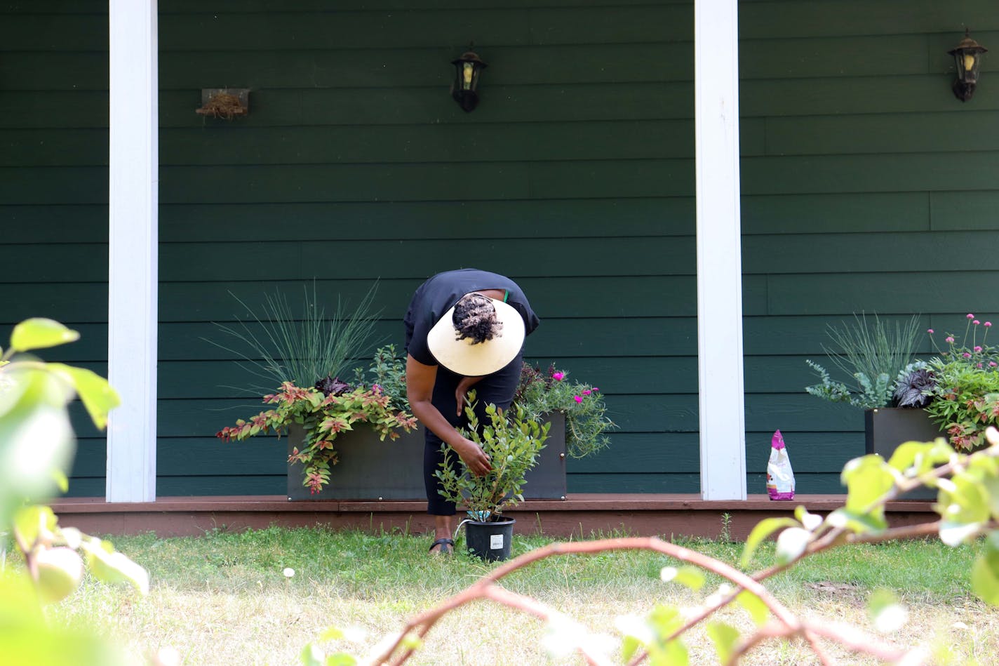 A woman with a sunhat crouches down to tend to potted plants on a deck. Behind her is the dark green lap siding of her rural home.