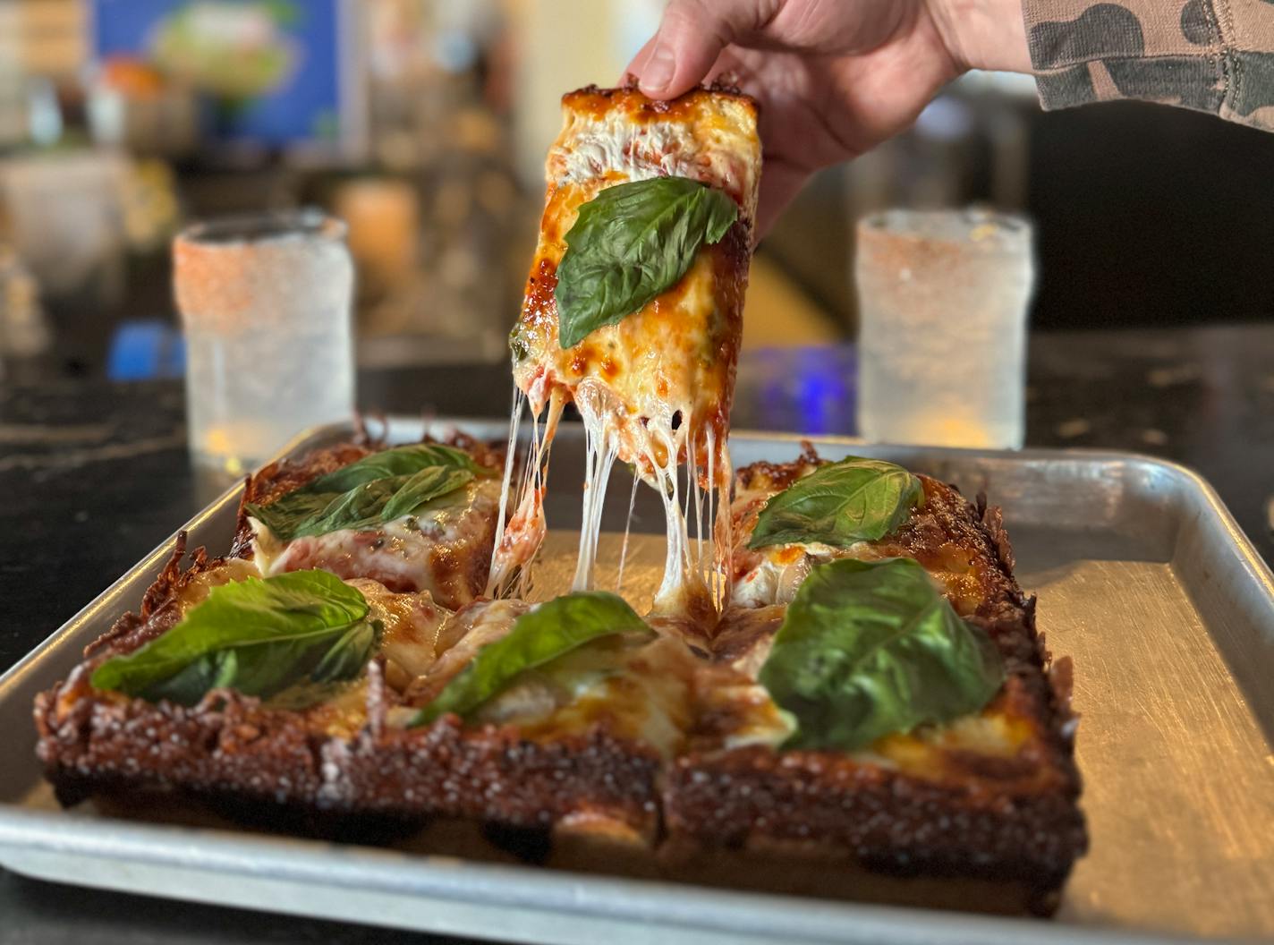 A hand pulls up a large, thick, gooey square of pizza from a pan of Wrecktangle pizza inside the taproom at Wild State Cider.