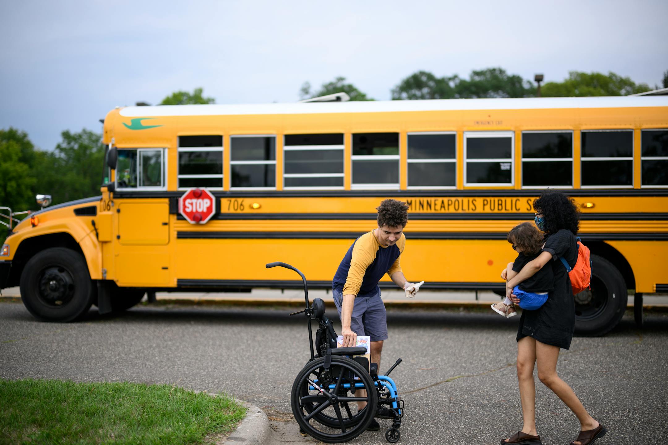 Sienna Schuth carries her son, Meru, as co-parent Dane Johnson prepares Meru's wheelchair.