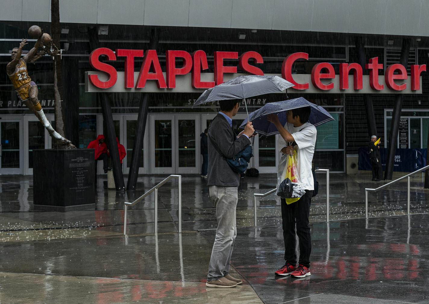 Tourist stand under umbrellas in rain outside Staples Center, home to hockey's Los Angeles Kings and basketball's Los Angeles Lakers, Clippers and Sparks, in Los Angeles on Thursday, March 12, 2020. Officials have banned large gatherings and events in Los Angeles to try to stop the spread of the new coronavirus. (AP Photo/Damian Dovarganes)